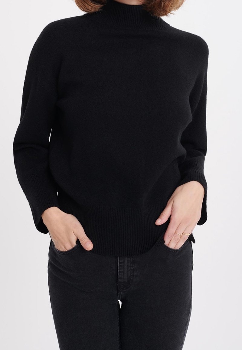 Czarny sweter z golfem o luźnym kroju, z prążkowanymi mankietami i dołem. Materiał wydaje się miękki i teksturowany. Noszony z czarnymi jeansami.