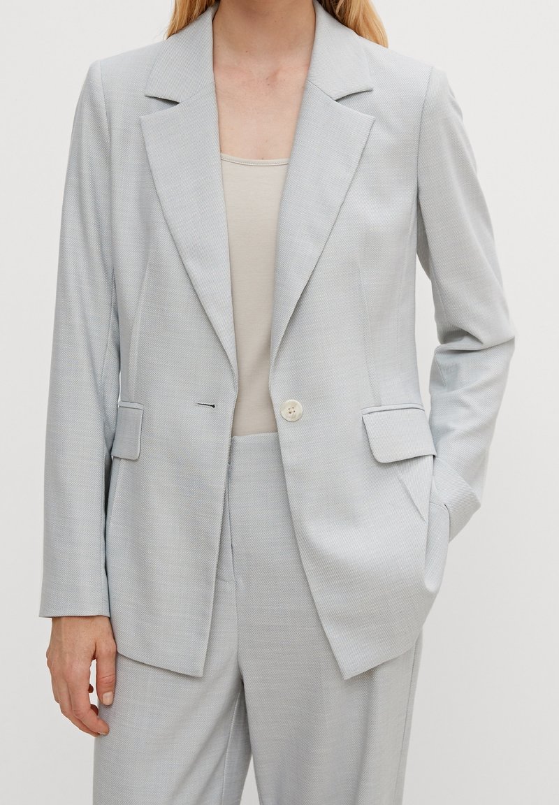 Femme portant un blazer gris clair sur mesure et un pantalon assorti avec un haut beige en dessous, une main dans la poche, debout devant un fond uni.