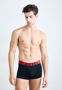 Schwarze Boxer-Briefs mit einem roten elastischen Bund, der das 'DIESEL'-Logo trägt. Glattes Material und eng anliegendes Design. Modell steht in neutraler Pose.