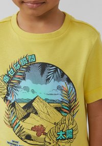 Geel katoenen T-shirt met een circulaire graphic van bergen, een voertuig en tropische bladeren, met tekst in het Japans en Engels.