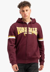 Bordový zipsový mikina s bielymi a žltými pruhmi na rukávoch, veľké logo "LONSDALE" v žltej farbe, predné vrecká a rebrovaný lem.