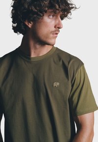 Olijfgroene katoenen T-shirt met korte mouwen, voorzien van een klein geborduurd olifantenlogo op de linkerkant van de borst. Glad textuur, ronde hals.