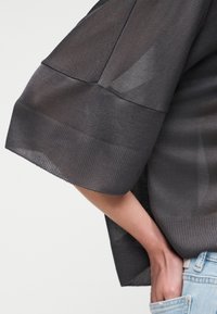 Personne portant un haut ample gris foncé texturé avec des manches larges, main glissée dans la poche d'un jean en denim bleu clair.