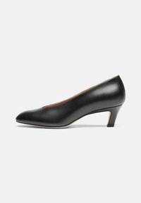 Zapato de charol negro con punta afilada, tacón bajo tipo kitten heel, textura suave y diseño minimalista. Presenta un borde sutil en la suela.