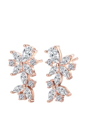 Mujer con pendientes de diamantes en forma de flor, tocándose un lóbulo de la oreja con dedos manicuraos, cabello recogido con una cinta beige.