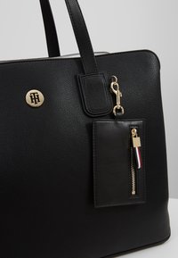 Sac à main en cuir noir avec double sangle, emblème logo doré, et pochette porte-clés en cuir noir attachée avec fermeture éclair dorée et ruban rouge-blanc-bleu.