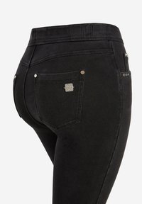 Jeans elásticos negros con un talle medio y una silueta ceñida. Las características incluyen dos bolsillos traseros, herrajes metálicos y costuras sutiles.