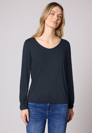 Kobieta o długich blond włosach, ubrana w czarną koszulę z długim rękawem i niebieskie dżinsy, na jasnoszarym, jednolitym tle.