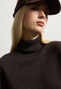 Maglione a collo alto di colore marrone, realizzato in morbido materiale a maglia con dettagli a costine. Un berretto marrone scuro completa il look, caratterizzato da una visiera strutturata.