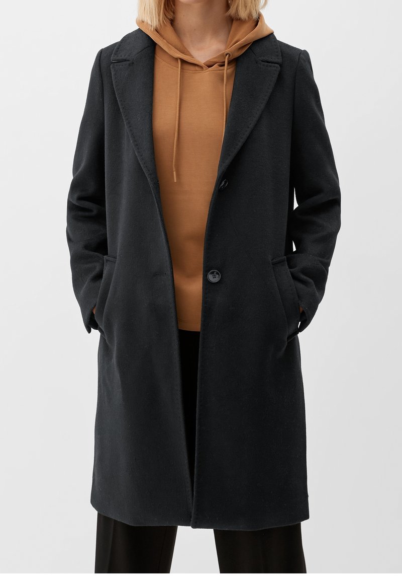 Svart ullblandad kappa med strukturerad design, spetsiga slag, framknäppning och sidofickor, bärs över en brun huvtröja.