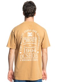 Beige cotton t-shirt featuring white text and graphics on the back, including "DC" logo and "DESTROY & CREATE" branding. Short sleeves.