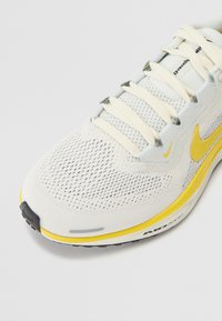 Chaussure de course Nike en mesh gris clair avec des accents jaunes. Présente un bout arrondi, une languette rembourrée et des lacets, ainsi qu'une semelle et un talon texturés.