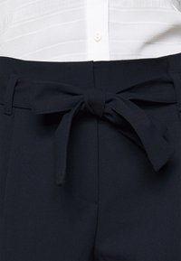 Marinblå byxor med en slät yta, prydda med en knuten rosett vid midjan och diskreta veckdetaljer fram, designade för en skräddarsydd passform.