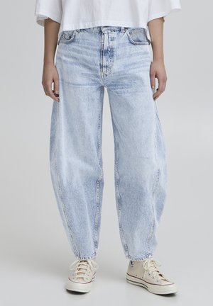 Jeans de denim azul claro con un corte relajado, que presentan una cintura alta, piernas rectas y un desvanecimiento sutil. Combinados con zapatillas de lona beige.