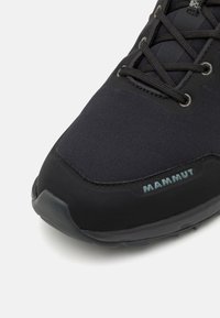 Chaussure de randonnée noire avec un dessus texturé, un capuchon en caoutchouc à l'avant et des lacets ; logo "MAMMUT" en bleu clair sur le côté. Vue en gros plan.