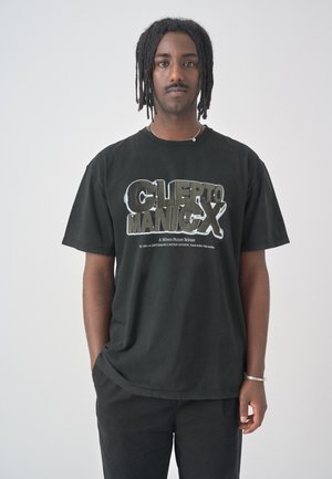Junger Mann mit geflochtenem Haar trägt ein schwarzes T-Shirt mit Grafik und Hose, steht vor einem hellen einfarbigen Hintergrund und schaut mit neutralem Gesichtsausdruck nach vorne.