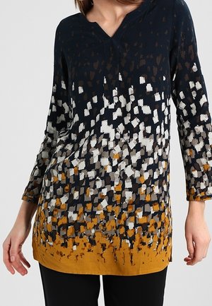 Kobieta ubrana w długą tunikę z rozproszonym wzorem kwadratów w kolorach czarnym, białym, szarym i musztardowym, w połączeniu z czarnymi spodniami.