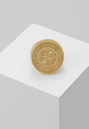 Złoty okrągły pierścień z wygrawerowanym tekstem "GUESS EST 1981 LOS ANGELES" i splecionym czteroliterowym logo na białym sześcianie.