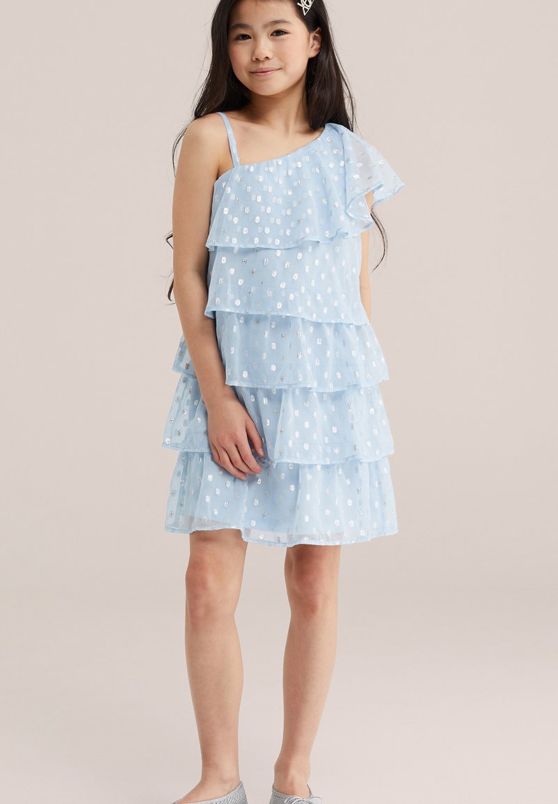 Jeune fille aux longs cheveux foncés portant une robe bleu clair à volants avec des points argentés et une barrette, se tenant devant un fond uni.