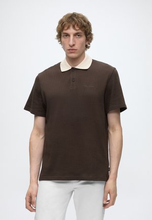 Young man with curly hair wearing a dark brown textured polo shirt with a white collar and white pants, standing against a plain light background.