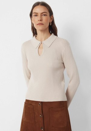 Frau trägt ein beiges, geripptes Langarmshirt mit Kragen und Schlüsselloch-Ausschnitt, kombiniert mit einem braunen Rock mit Knopfleiste und Taschen.