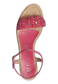 Pink flower-embellished sandal with a flat, tan sole and adjustable ankle strap; features a smooth, textured footbed with metallic logo.