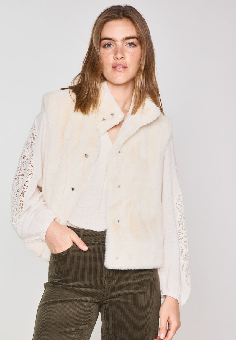 Gilet in pelliccia sintetica color crema con una texture morbida, caratterizzato da un colletto a punta e bottoni a pressione. Il modello indossa una maglietta bianca e pantaloni in velluto a coste verde oliva.