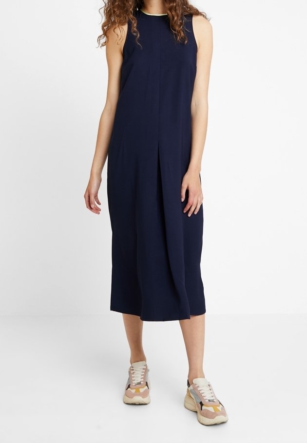 Femme portant une robe midi bleu marine sans manches avec une fente latérale et des baskets multicolores, debout devant un fond blanc.