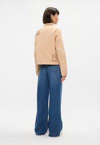 Μπεζ βαμβακερό μπουφάν με γιακά, κεντητό φύλλο στην πλάτη και λεπτομέρεια λογότυπου στο πλάι. Συνδυασμένο με τζιν παντελόνι σε μπλε χρώμα και λευκά αθλητικά παπούτσια.