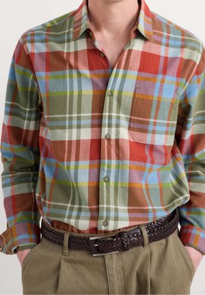 Homme portant une chemise à carreaux colorée boutonnée rentrée dans un pantalon kaki avec une ceinture en cuir tressé brun foncé. Mains dans les poches.