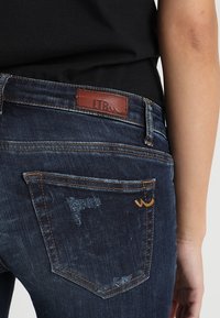 Vaqueros de mezclilla oscura con un parche de cuero con el logo LTB en la cintura, costuras en contraste y detalles desgastados en los bolsillos traseros.