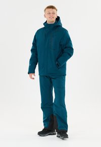 Mann in teal-farbener wasserdichter Jacke mit Kapuze und passender Hose, der mit einer Hand in der Tasche steht und lächelnd zur Seite blickt.
