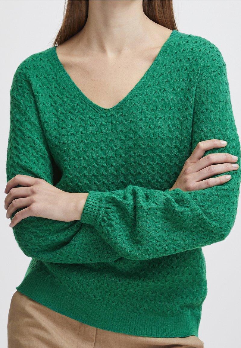 Pull vert texturé à col en V porté par une personne les bras croisés, assorti à un pantalon beige, sur fond blanc.