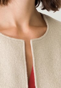 Beige, gestructureerde, wolblend jas met een ronde hals en open voorkant. Toont een rode top onder de kraag van de jas.