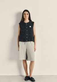Donkergrijs mouwloos gebreid topje met een lichte textuur, voorzien van een rij knopen en een label; gecombineerd met lichtgrijze shorts en zwarte instappers.