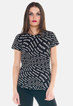 Jeune femme aux cheveux bruns lisses porte un t-shirt noir recouvert de texte blanc « STAY ACTIVE » en motifs diagonaux.
