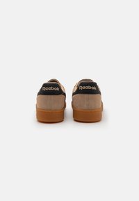 Baskets en daim beige avec un contrefort de talon noir portant la marque Reebok. Semelle en caoutchouc marron avec des détails texturés.