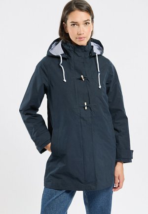 Femme portant un imperméable à capuche bleu marine avec cordons blancs et boutons à bascule, posant avec une main dans la poche et un jean bleu.