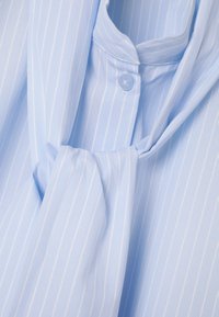 Light blue striped blouse with a soft, smooth texture. Features a button closure and a tied scarf detail at the neckline.