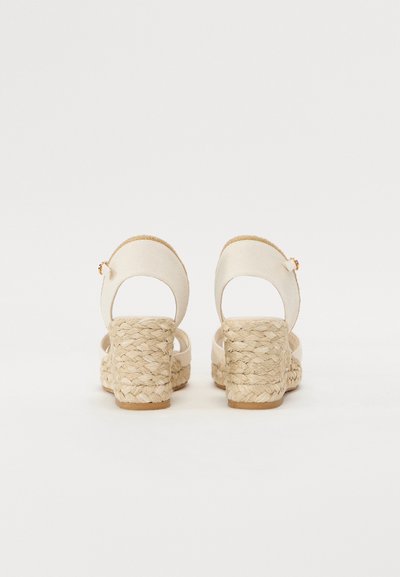 Kaksi taaksepäin näkyvää viistokorkoista sandaalia, joissa on beige kangas päällinen, kudottu juuttikorko ja ohut nilkkaremmi kultasävytetyllä soljella.