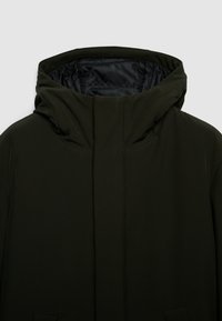 Giacca con cappuccio verde scuro con collo alto, esterno liscio, cuciture discrete e due tasche frontali. Interno foderato in nero.