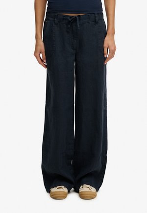 Persona vistiendo pantalones negros de corte holgado con bolsillos delanteros y cintura con cordón, combinados con zapatos beige con cordones.