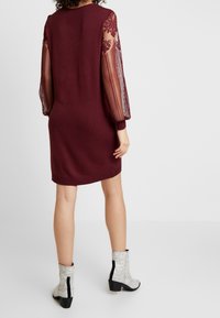 Donna che indossa un abito di maglia bordeaux con maniche a palloncino in pizzo trasparente, abbinato a stivaletti alla caviglia con motivo in pelle di serpente bianco, in piedi su un pavimento bianco.