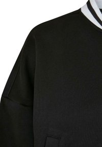 Musta collegepaita, jossa on teksturoidusta kankaasta valmistettu pinta. Paidassa on pyöreä pääntie, jossa on valkoiset ja mustat raidat, sekä raglan-hihat.