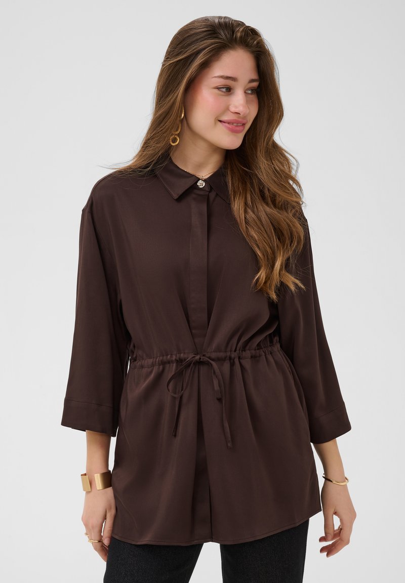 Femme aux longs cheveux ondulés portant une robe-chemise marron foncé boutonnée avec une taille à cordon et des bijoux en or, souriante et regardant vers la droite.