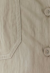 Tejido beige con costuras anguladas blancas y dos botones beige cosidos con hilo beige en el lado derecho.