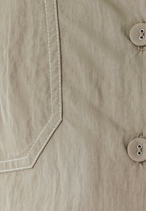 Bežinis audinys su balta kampine siūle ir dviem bežiniais mygtukais, siūtais bežiniu siūlu dešinėje pusėje.