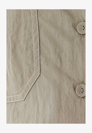 Bežinis audinys su balta kampine siūle ir dviem bežiniais mygtukais, siūtais bežiniu siūlu dešinėje pusėje.