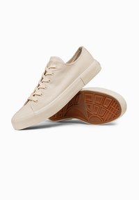 Paire de baskets basses en toile beige avec lacets beiges et semelles en caoutchouc, une chaussure reposant sur la semelle de l’autre.