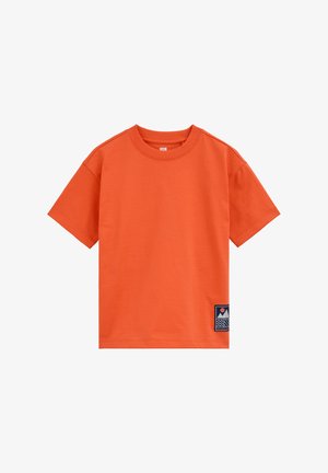 Narančasta majica kratkih rukava s okruglim izrezom i malim pravokutnim zakrpom s motivom planine i sunca blizu donjeg ruba s desne strane.